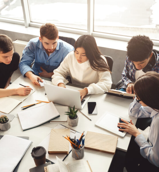 Eine Gruppe von Studenten arbeitet gemeinsam an einem Tisch mit Laptops. Sie sind in eine konzentrierte Teamarbeit vertieft und umgeben von Schreibmaterialien wie Stifte, Notizbücher und Ordner. Die Szene vermittelt ein produktives Lernumfeld mit moderner Technologie.