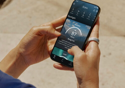 Ein Mensch hält ein Smartphone in beiden Händen und nutzt eine Gesundheits-Tracking-App. Am rechten Zeigefinger trägt die Person einen silbernen Smart Ring. Die App auf dem Bildschirm zeigt eine "Readiness"-Bewertung von 87 an, begleitet von weiteren Gesundheitsmetriken. Der Hintergrund ist leicht verschwommen und zeigt einen sonnigen Außenbereich mit einem hellen Bodenbelag. Die Person trägt ein blaues Oberteil.