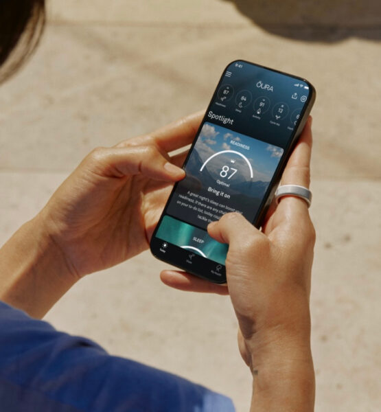 Ein Mensch hält ein Smartphone in beiden Händen und nutzt eine Gesundheits-Tracking-App. Am rechten Zeigefinger trägt die Person einen silbernen Smart Ring. Die App auf dem Bildschirm zeigt eine "Readiness"-Bewertung von 87 an, begleitet von weiteren Gesundheitsmetriken. Der Hintergrund ist leicht verschwommen und zeigt einen sonnigen Außenbereich mit einem hellen Bodenbelag. Die Person trägt ein blaues Oberteil.