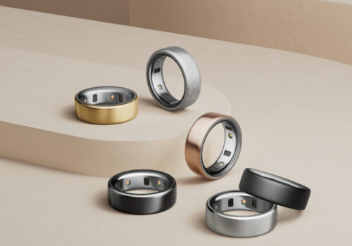 Ein stilvoll arrangiertes Set aus sechs modernen Smart-Ringen in verschiedenen Metallfarben – darunter Gold, Schwarz, Silber, Roségold und gebürstetes Grau – auf einer minimalistischen beigen Stufenplattform. Die Ringe sind mit eingebauten Sensoren auf der Innenseite ausgestattet und reflektieren einen eleganten, technologischen Look. Die Komposition betont die luxuriöse Verarbeitung und die Vielseitigkeit der Smart-Ringe als modische Wearables.
