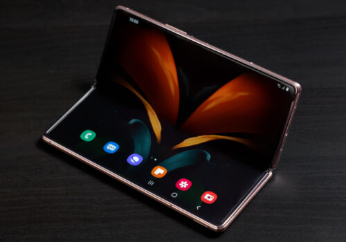 Ein modernes faltbares Smartphone liegt auf einer dunklen Oberfläche und ist halb aufgeklappt. Der große, flexible Bildschirm zeigt eine elegante, abstrakte Hintergrundgrafik mit leuchtenden Farben. Die Benutzeroberfläche ist sichtbar, mit mehreren App-Symbolen am unteren Rand des Displays. Das Gerät hat ein schlankes, hochwertiges Design mit metallischem Rahmen und nahtlosem Displayübergang.
