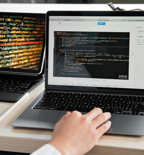 Auf einem Tisch stehen ein Tablet und ein Laptop. Auf den Bildschirmen ist Code zu sehen und eine Hand beient grade die Tastatur des Laptops.