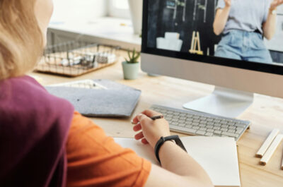 Eine Frau sitzt mit Stift und Heft vor ihrem Computerbildschirm. Auf dem Bildschirm ist eine weitere Frau, eine Lehrperson, vor einer Tafel zu sehen.
