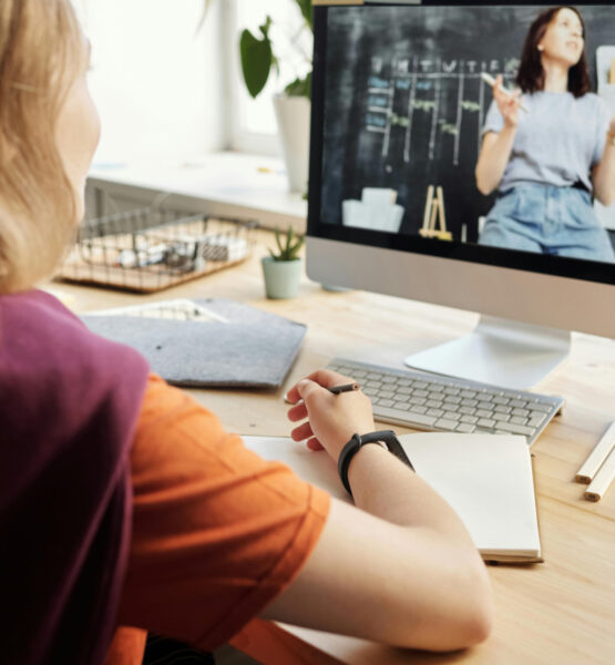 Eine Frau sitzt mit Stift und Heft vor ihrem Computerbildschirm. Auf dem Bildschirm ist eine weitere Frau, eine Lehrperson, vor einer Tafel zu sehen.