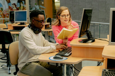 Ein Mann und eine Frau arbeiten gemeinsam an einem Computer. Der Mann tippt an der Tastatur während die Frau ihm mit der einen Hand ein Buch entgegenstreckt und mit der anderen etwas auf dem Bildschirm zeigt.