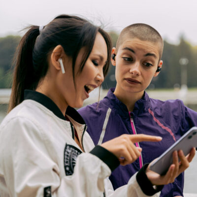 Zwei junge Frauen stehen im Freien an einem Gewässer und schauen gemeinsam auf ein Smartphone. Die Frau links lacht begeistert und zeigt mit dem Finger auf den Bildschirm, während die Frau rechts mit kurzem Haar und lila Sportjacke interessiert, aber etwas skeptisch auf das Gerät blickt. Beide tragen kabellose Ohrhörer. Im Hintergrund sind Bäume und ein Springbrunnen zu sehen.