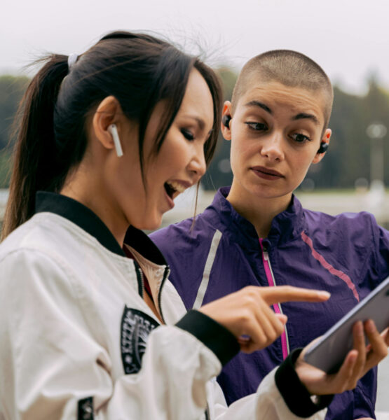 Zwei junge Frauen stehen im Freien an einem Gewässer und schauen gemeinsam auf ein Smartphone. Die Frau links lacht begeistert und zeigt mit dem Finger auf den Bildschirm, während die Frau rechts mit kurzem Haar und lila Sportjacke interessiert, aber etwas skeptisch auf das Gerät blickt. Beide tragen kabellose Ohrhörer. Im Hintergrund sind Bäume und ein Springbrunnen zu sehen.