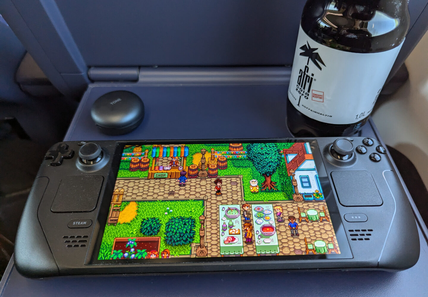 Steam Deck OLED und Getränk auf dem Tisch eines Zuges. Auf dem Gerät läuft gerade Stardew Valley.