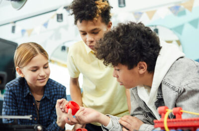 Drei Kinder beugen sich über einen Tisch und interagieren miteinander in einer Gruppenarbeit. Auf dem Tisch sind Robotikmaterialien.