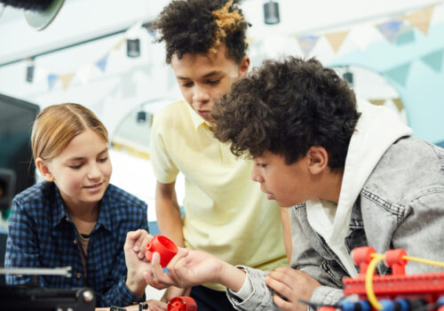 Drei Kinder beugen sich über einen Tisch und interagieren miteinander in einer Gruppenarbeit. Auf dem Tisch sind Robotikmaterialien.