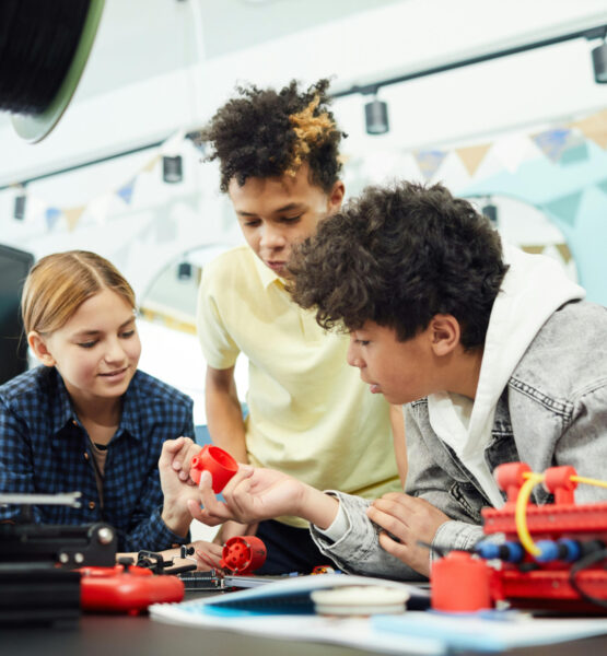 Drei Kinder beugen sich über einen Tisch und interagieren miteinander in einer Gruppenarbeit. Auf dem Tisch sind Robotikmaterialien.