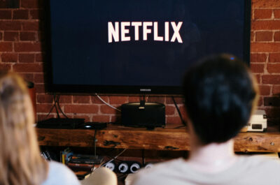 Auf einem Fernseher ist das Netflixlogo zu sehen. Davor sitzen, etwas unschärfer, zwei Personen auf einer Couch.