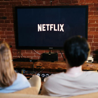 Auf einem Fernseher ist das Netflixlogo zu sehen. Davor sitzen, etwas unschärfer, zwei Personen auf einer Couch.