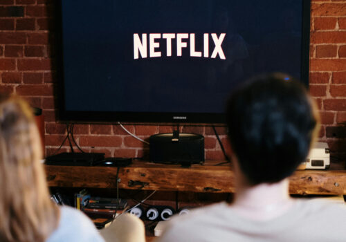 Auf einem Fernseher ist das Netflixlogo zu sehen. Davor sitzen, etwas unschärfer, zwei Personen auf einer Couch.