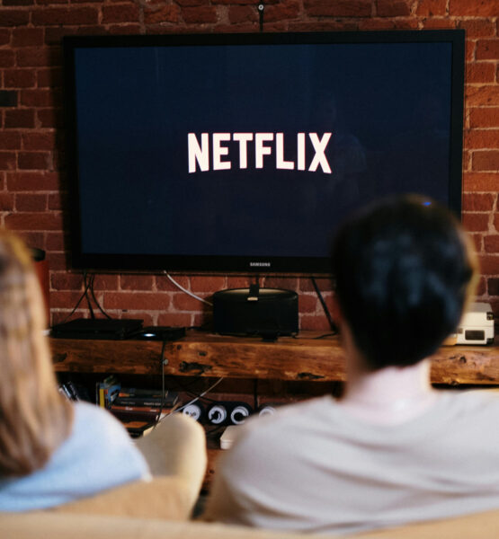 Auf einem Fernseher ist das Netflixlogo zu sehen. Davor sitzen, etwas unschärfer, zwei Personen auf einer Couch.