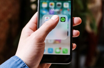 Auf einem IPhone ist ein Ordner mit dem Titel Musik geöffnet. Ein Daumen verdeckt einige App-Icons, doch Spotify ist zu sehen.