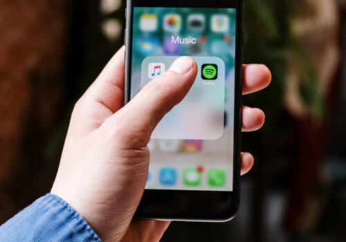 Auf einem IPhone ist ein Ordner mit dem Titel Musik geöffnet. Ein Daumen verdeckt einige App-Icons, doch Spotify ist zu sehen.