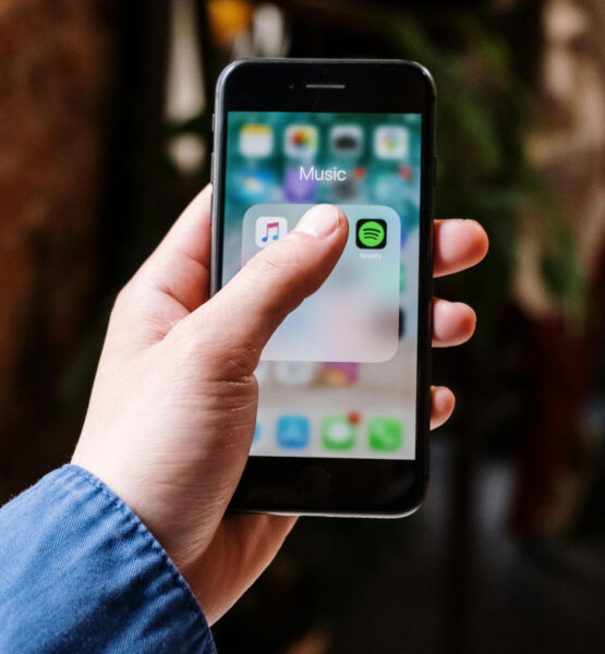 Auf einem IPhone ist ein Ordner mit dem Titel Musik geöffnet. Ein Daumen verdeckt einige App-Icons, doch Spotify ist zu sehen.
