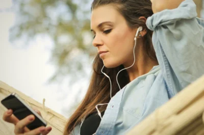 Eine junge Frau sitzt entspannt in einer Hängematte im Freien, trägt Kopfhörer und schaut auf ihr Smartphone. Sie wirkt ruhig und in ihre Musik oder einen Podcast vertieft.