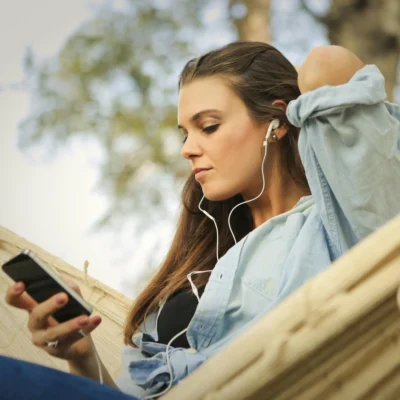 Eine junge Frau sitzt entspannt in einer Hängematte im Freien, trägt Kopfhörer und schaut auf ihr Smartphone. Sie wirkt ruhig und in ihre Musik oder einen Podcast vertieft.