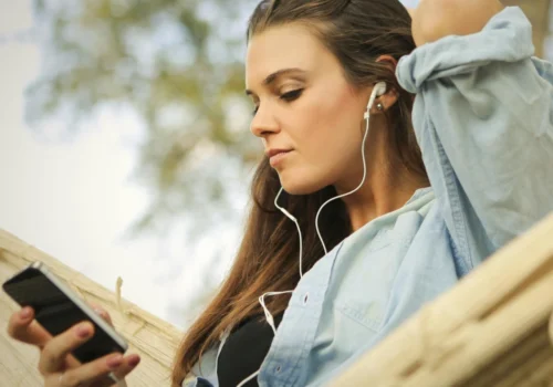 Eine junge Frau sitzt entspannt in einer Hängematte im Freien, trägt Kopfhörer und schaut auf ihr Smartphone. Sie wirkt ruhig und in ihre Musik oder einen Podcast vertieft.