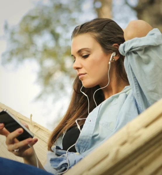 Eine junge Frau sitzt entspannt in einer Hängematte im Freien, trägt Kopfhörer und schaut auf ihr Smartphone. Sie wirkt ruhig und in ihre Musik oder einen Podcast vertieft.