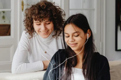 Zwei junge Frauen sitzen auf einem Sofa und hören gemeinsam über Kopfhörer einen Podcast. Die eine lehnt sich lächelnd über die Rückenlehne, während die andere entspannt zuhört – eine gemütliche, freundliche Hörsituation im Wohnzimmer.
