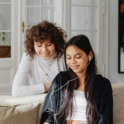 Zwei junge Frauen sitzen auf einem Sofa und hören gemeinsam über Kopfhörer einen Podcast. Die eine lehnt sich lächelnd über die Rückenlehne, während die andere entspannt zuhört – eine gemütliche, freundliche Hörsituation im Wohnzimmer.