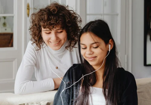 Zwei junge Frauen sitzen auf einem Sofa und hören gemeinsam über Kopfhörer einen Podcast. Die eine lehnt sich lächelnd über die Rückenlehne, während die andere entspannt zuhört – eine gemütliche, freundliche Hörsituation im Wohnzimmer.