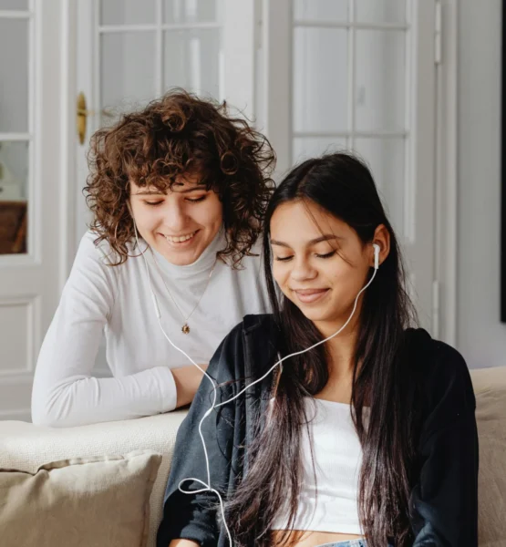 Zwei junge Frauen sitzen auf einem Sofa und hören gemeinsam über Kopfhörer einen Podcast. Die eine lehnt sich lächelnd über die Rückenlehne, während die andere entspannt zuhört – eine gemütliche, freundliche Hörsituation im Wohnzimmer.