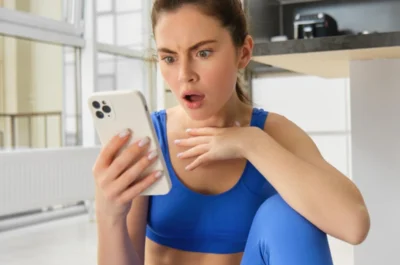 Eine Junge Influencerin in blauer Sportkleidung sitzt auf dem Boden einer hellen Küche und schaut überrascht auf ihr Smartphone, eine Hand vor der Brust, mit weit geöffneten Augen und leicht geöffnetem Mund.