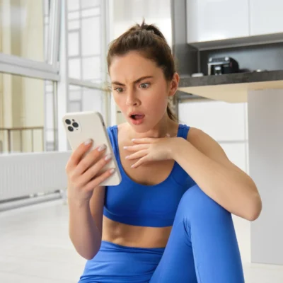 Eine Junge Influencerin in blauer Sportkleidung sitzt auf dem Boden einer hellen Küche und schaut überrascht auf ihr Smartphone, eine Hand vor der Brust, mit weit geöffneten Augen und leicht geöffnetem Mund.