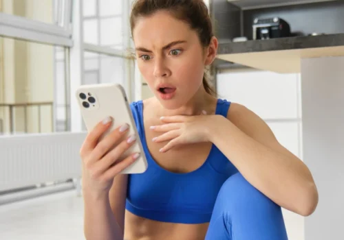 Eine Junge Influencerin in blauer Sportkleidung sitzt auf dem Boden einer hellen Küche und schaut überrascht auf ihr Smartphone, eine Hand vor der Brust, mit weit geöffneten Augen und leicht geöffnetem Mund.