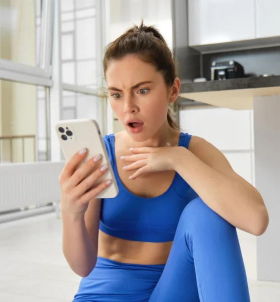 Eine Junge Influencerin in blauer Sportkleidung sitzt auf dem Boden einer hellen Küche und schaut überrascht auf ihr Smartphone, eine Hand vor der Brust, mit weit geöffneten Augen und leicht geöffnetem Mund.