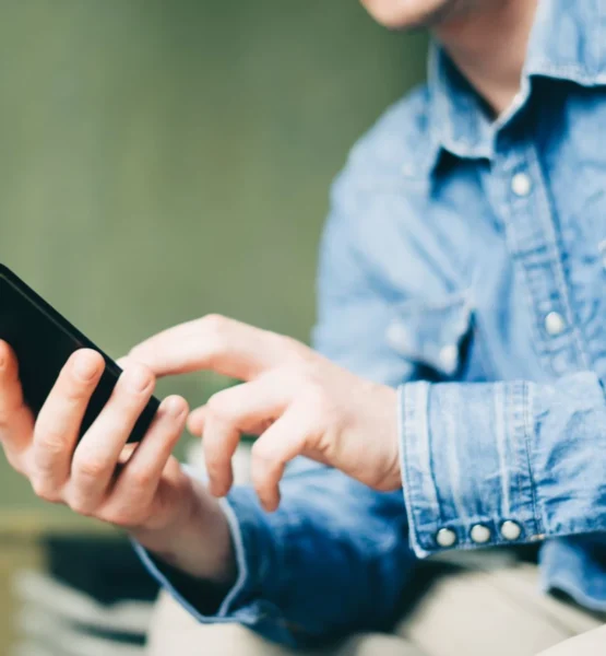 Eine Person sitzt draußen mit seinem Smartphone in der Hand und schaut sich auf diesem etwas an.