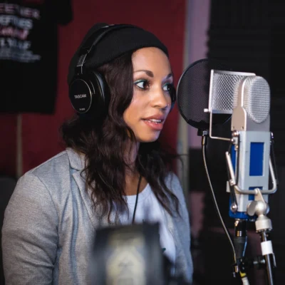 Eine Frau steht mit Kopfhörer vor einem Mikrofon im Aufnahmestudio.