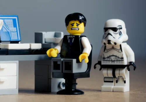 Ein LEGO-Männchen im Anzug sitzt gestresst an einem Schreibtisch vor einem Computer, während hinter ihm eine LEGO-Figur eines Stormtroopers steht.