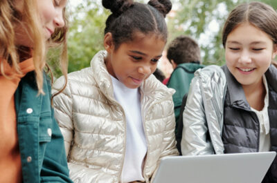 Drei junge Mädchen sitzen nebeneinander und schauen gemeinsam auf den Laptop auf dem Schoß des Mädchens in der Mitte