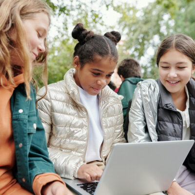 Drei junge Mädchen sitzen nebeneinander und schauen gemeinsam auf den Laptop auf dem Schoß des Mädchens in der Mitte