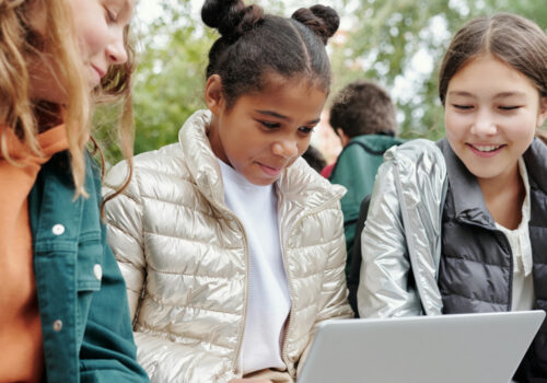 Drei junge Mädchen sitzen nebeneinander und schauen gemeinsam auf den Laptop auf dem Schoß des Mädchens in der Mitte
