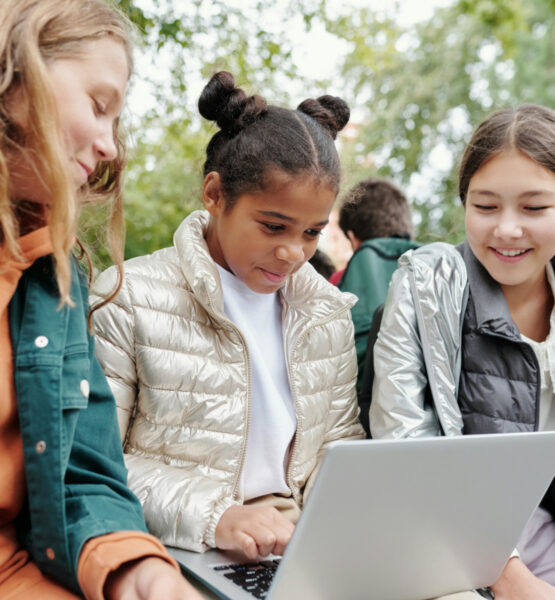 Drei junge Mädchen sitzen nebeneinander und schauen gemeinsam auf den Laptop auf dem Schoß des Mädchens in der Mitte