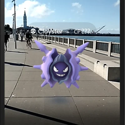 Ein Screenshot aus Pokémon GO zeigt ein lilafarbenes Pokémon (Cloyster), das scheinbar auf einem Gehweg an einer Uferpromenade schwebt. Im Hintergrund sind ein Geländer, Wasser, ein Fahrradfahrer und ein Turm zu sehen, während die reale Umgebung mit der Augmented-Reality-Darstellung kombiniert wird.