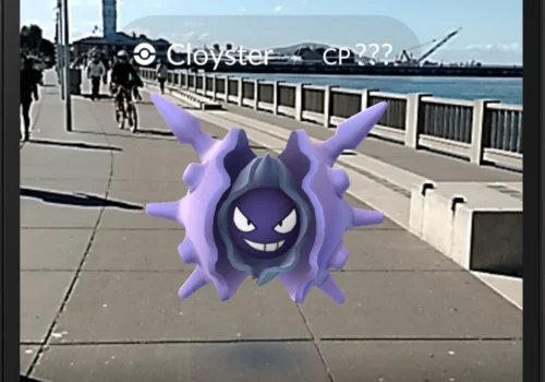 Ein Screenshot aus Pokémon GO zeigt ein lilafarbenes Pokémon (Cloyster), das scheinbar auf einem Gehweg an einer Uferpromenade schwebt. Im Hintergrund sind ein Geländer, Wasser, ein Fahrradfahrer und ein Turm zu sehen, während die reale Umgebung mit der Augmented-Reality-Darstellung kombiniert wird.
