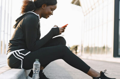 Frau im Trainingsanzug sitzt auf der Bordsteinkante und bedient ihr Smartphone