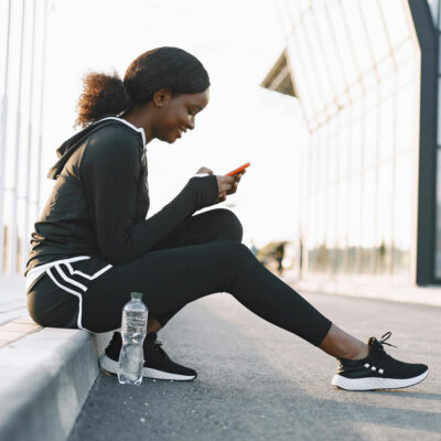 Frau im Trainingsanzug sitzt auf der Bordsteinkante und bedient ihr Smartphone