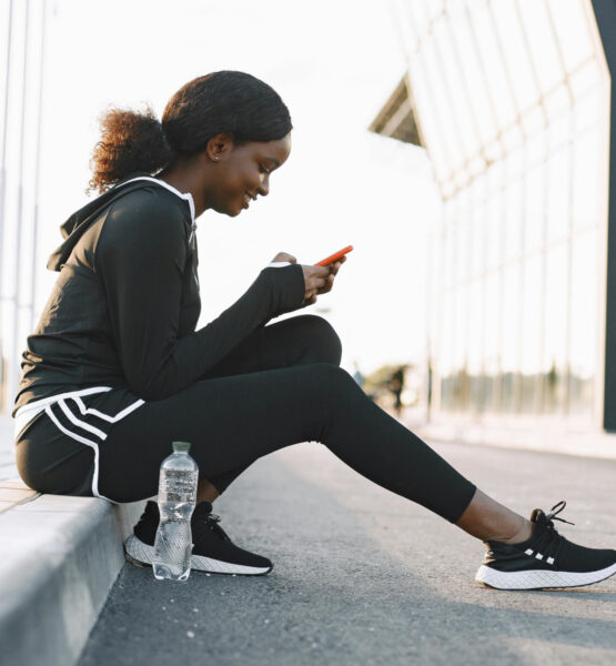 Frau im Trainingsanzug sitzt auf der Bordsteinkante und bedient ihr Smartphone