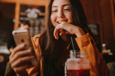 Eine Frau sitzt in einem Café und lacht während sie auf ihr Smartphone schaut.