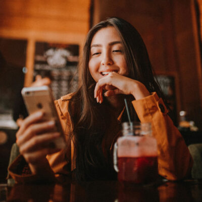 Eine Frau sitzt in einem Café und lacht während sie auf ihr Smartphone schaut.