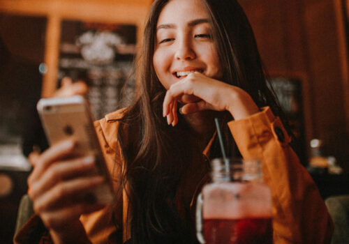 Eine Frau sitzt in einem Café und lacht während sie auf ihr Smartphone schaut.