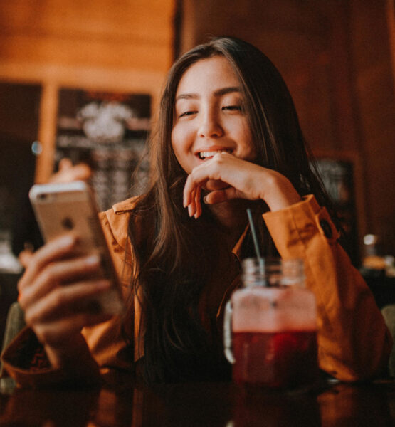 Eine Frau sitzt in einem Café und lacht während sie auf ihr Smartphone schaut.